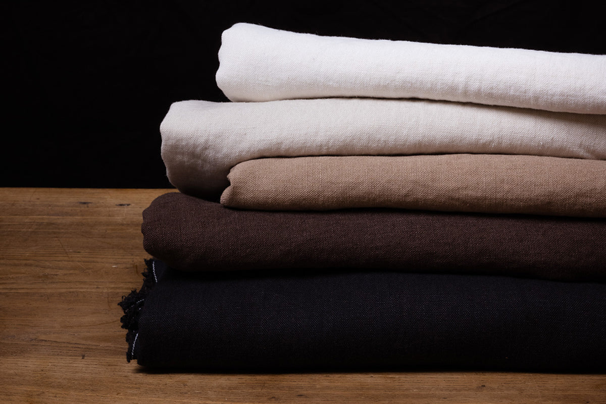 Stack of folded linen fabrics in white, beige, brown, and black on a wooden surface with a dark background.